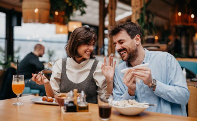Two Young Friends Meeting For Drinks And Food In Restaurant, laughing, and eating.
